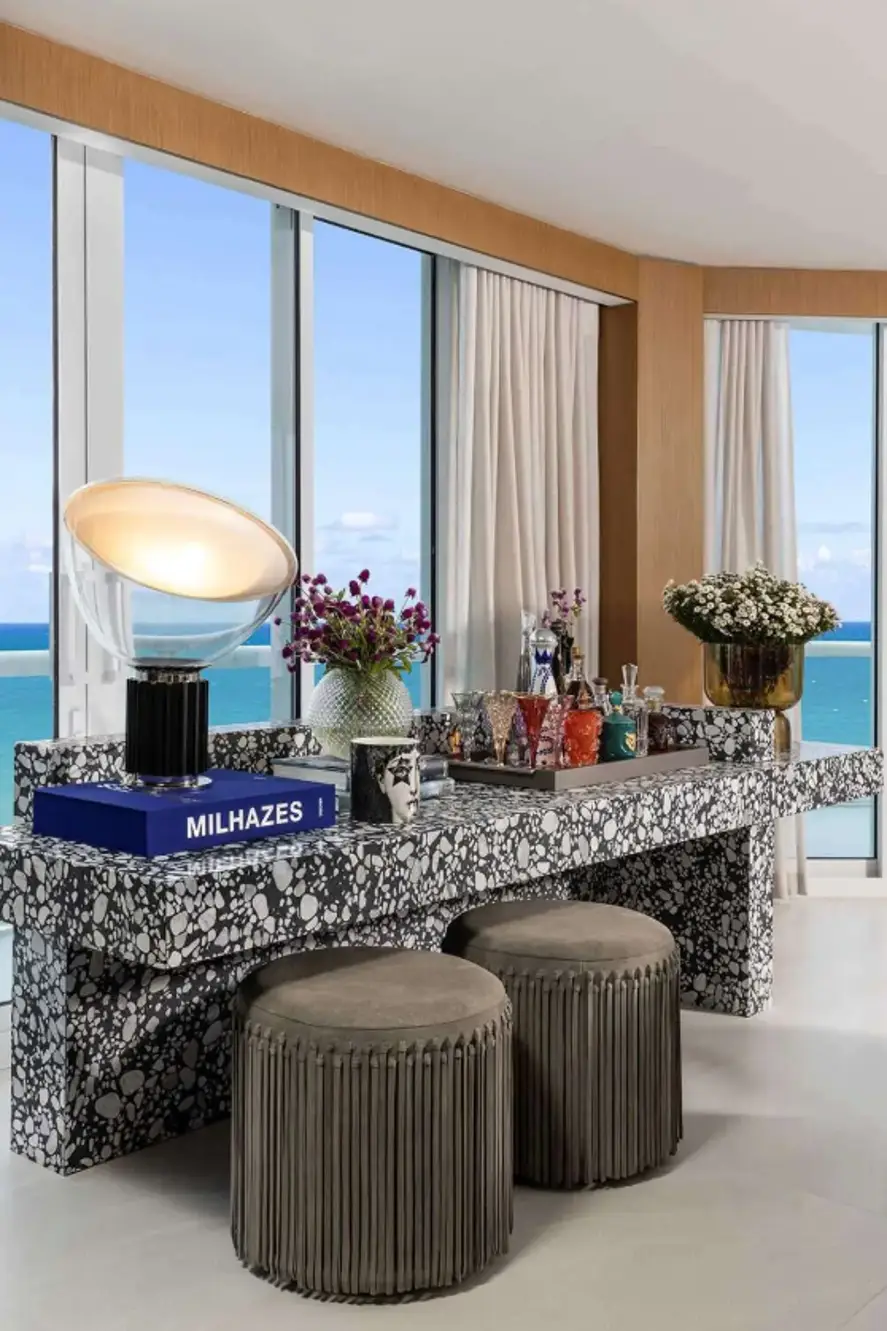 Modern terrazzo console table styled with eclectic glassware, sculptural decor, and floral arrangements, set against floor-to-ceiling windows with ocean views in a Bal Harbour condo.