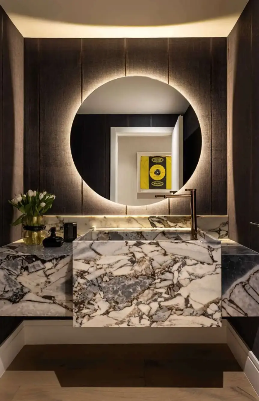 Luxurious powder room with dramatic stone vanity, backlit circular mirror, dark textured walls, and brass faucet accents.