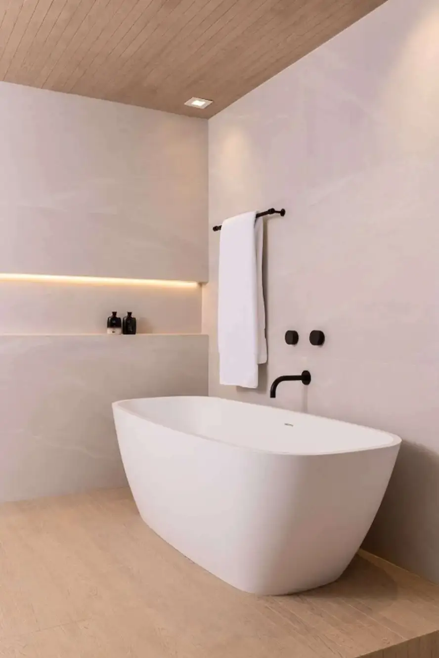 Freestanding white bathtub with matte black fixtures, set against a soft beige wall with integrated lighting and natural wood flooring and ceiling.