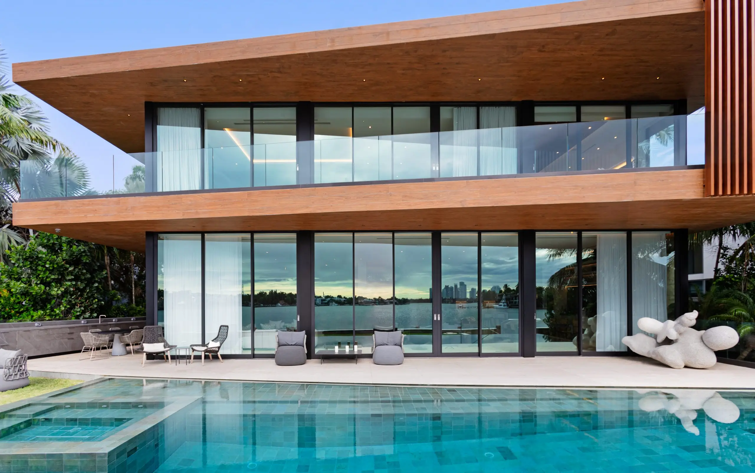 Modern Venetian Island home with waterfront views, glass walls, wood facade, and poolside sculpture overlooking Biscayne Bay.
