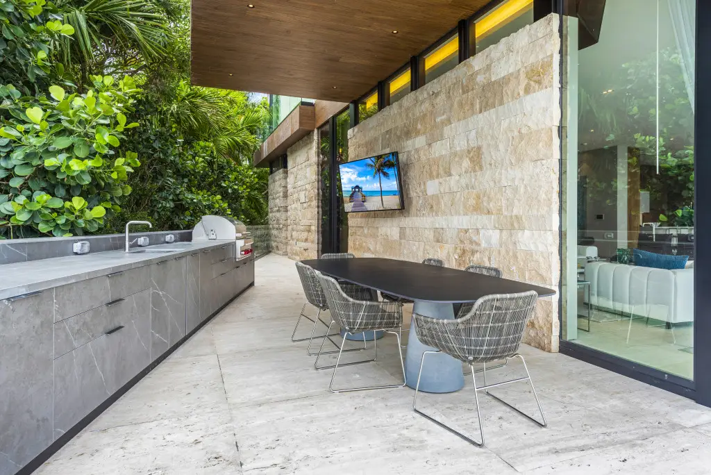 Modern outdoor dining area with stone wall, built-in grill, and wall-mounted TV in a Venetian Island luxury home