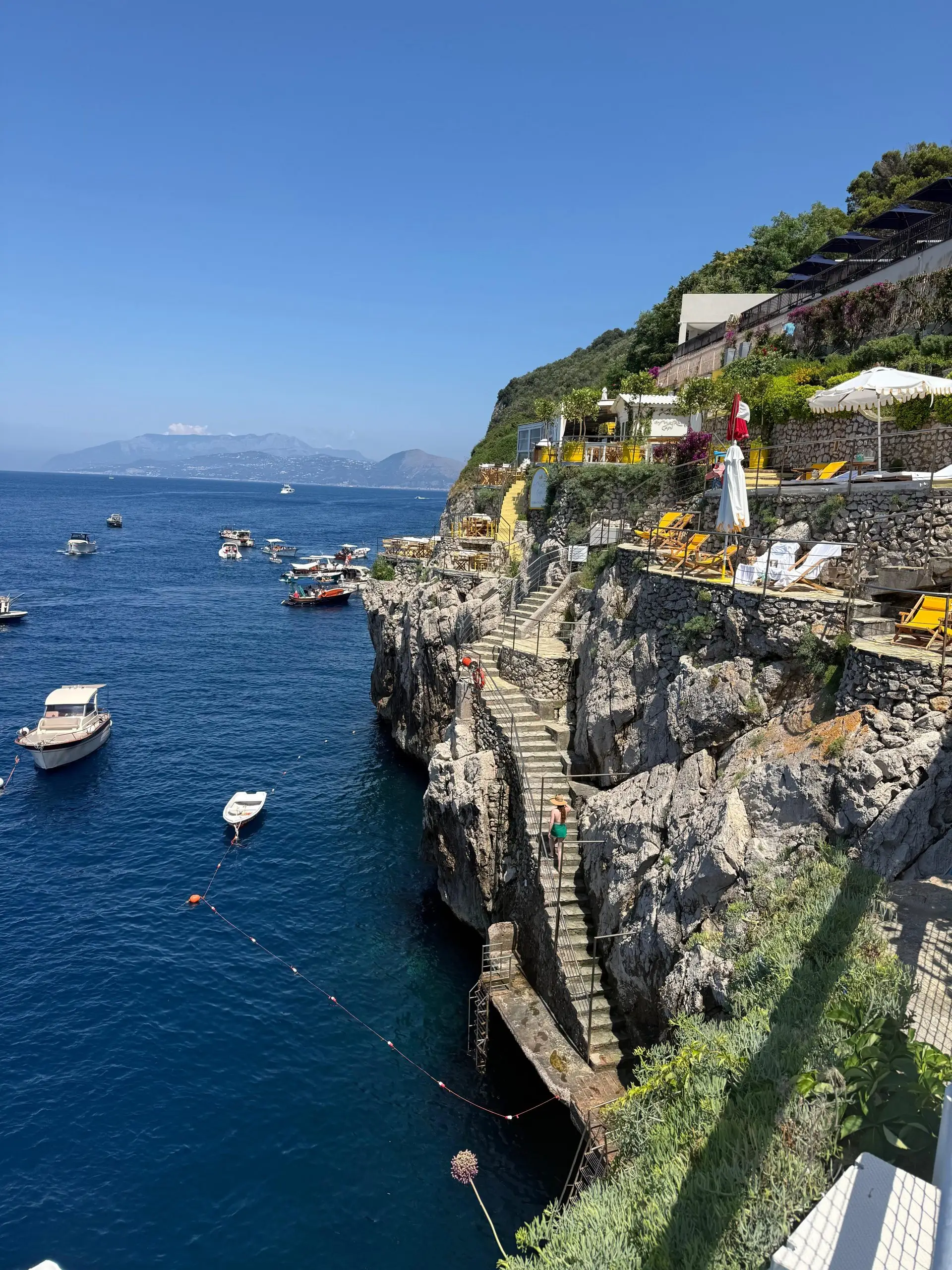 A vibrant summer scene in Italy showing how stone terraces and stairs are built into a cliffside, creating an outdoor space that overlooks the deep blue sea and nearby boats.